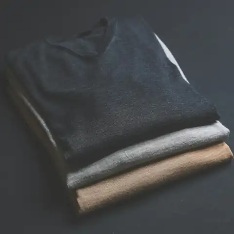 Três camisetas dobradas e empilhadas, em azul-marinho, cinza e bege, sobre fundo escuro