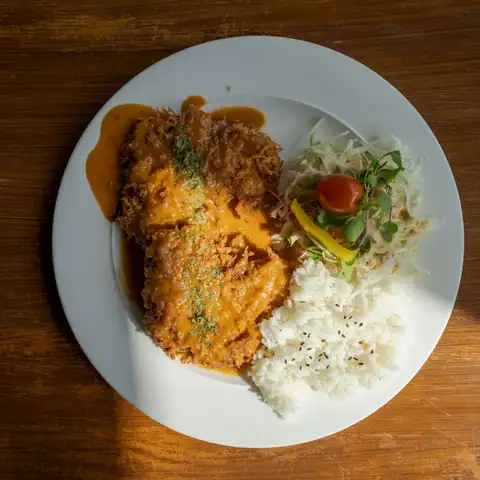 Filé de frango empanado com molho, arroz branco e salada num prato branco, visto de cima sobre uma mesa de madeira